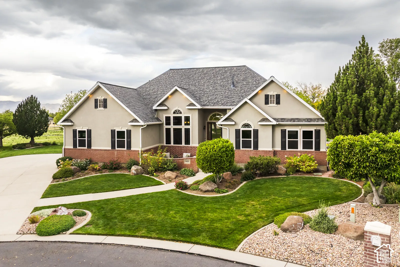 Stucco exterior with brick detailing, asphalt shingle roof, mature landscaping, expansive lawn, and a wide concrete driveway
