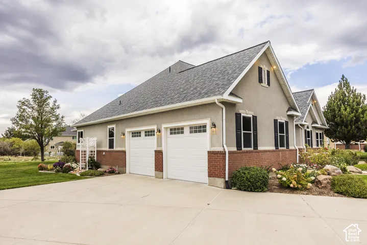 Stucco exterior with brick detailing, asphalt shingle roof, two-car garage with carriage-style doors, and established landscaping