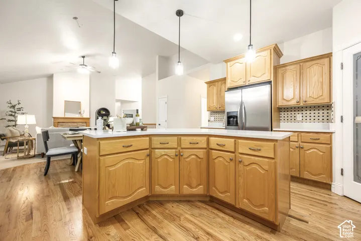Open-concept kitchen with wood-finish flooring, light wood cabinetry, stainless steel refrigerator, and a kitchen island with pendant lighting