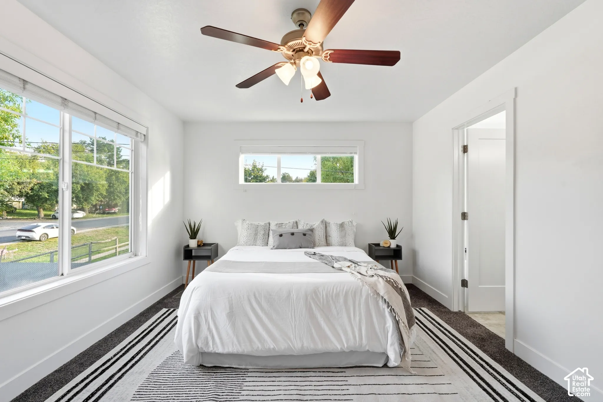 Bright white walls with a large window, a smaller transom window, a ceiling fan, and dark carpet flooring
