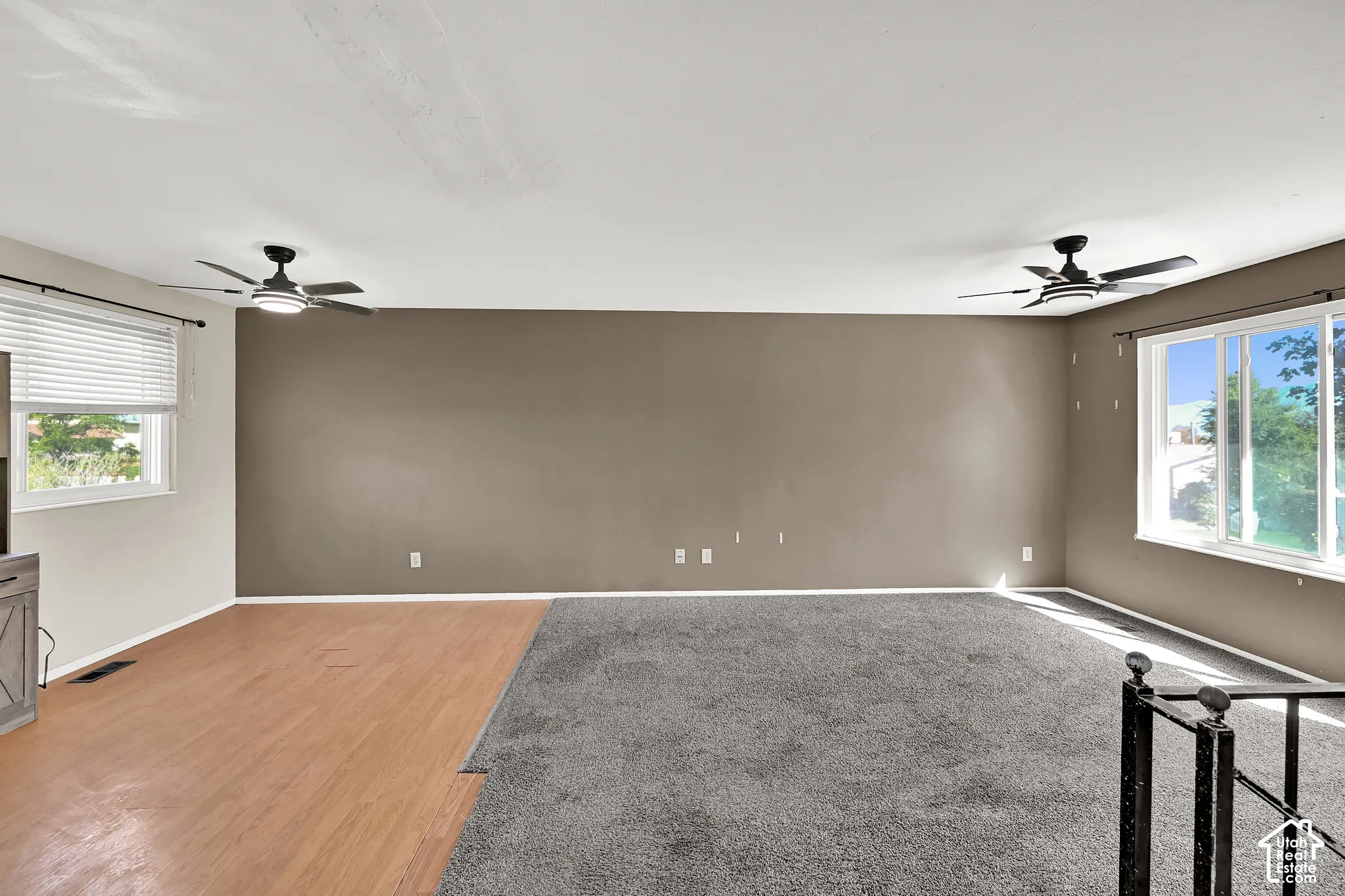 Room featuring mixed flooring with wood-finish and carpeted areas, two windows with blinds, and two ceiling fans with integrated lighting