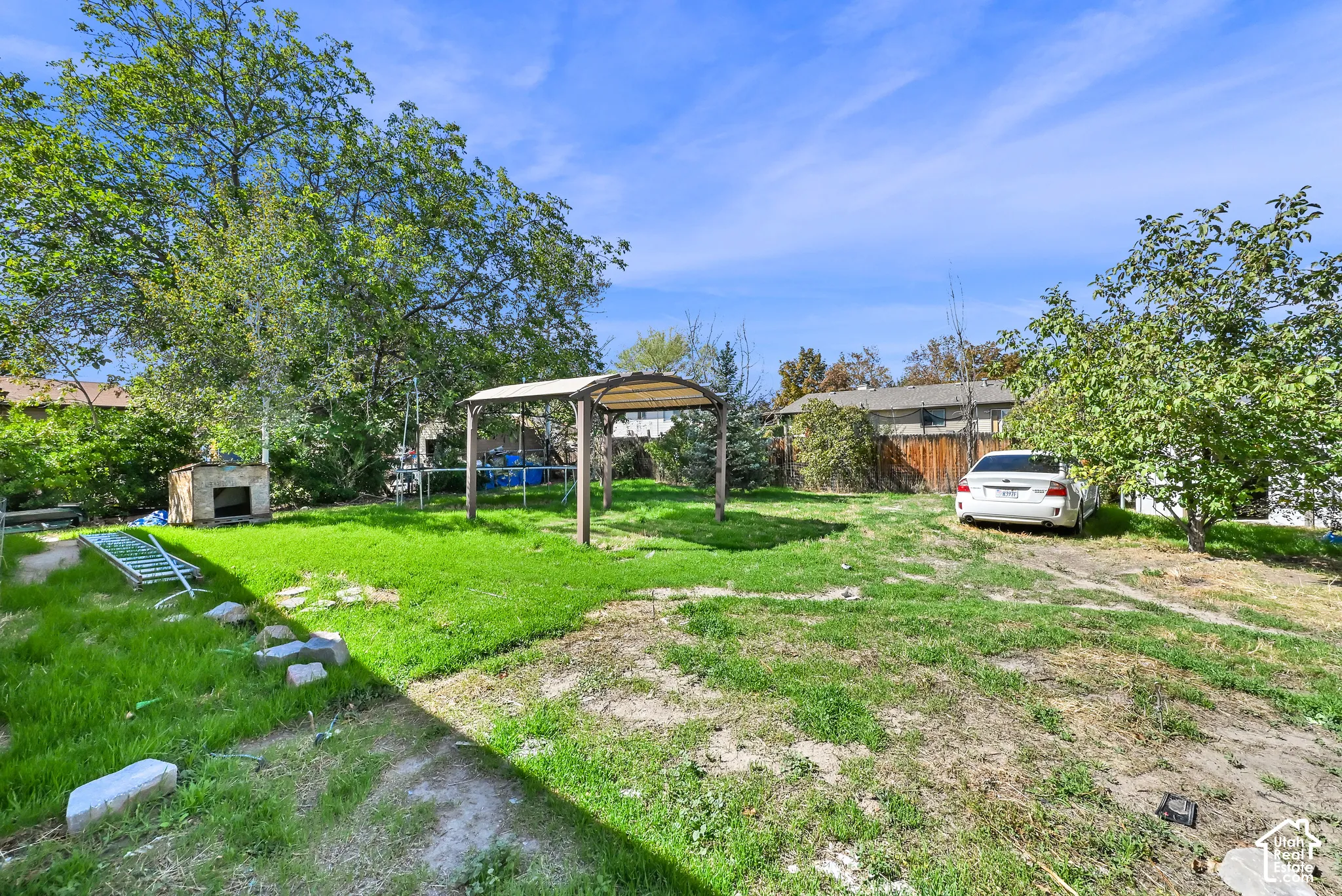 Expansive backyard featuring a partial grass lawn, mature trees, a built-in outdoor fireplace, and a standalone pergola