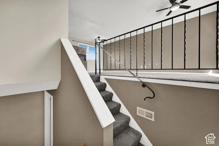 Carpeted staircase with white trim, leading to a lofted area featuring a black wrought iron railing and a ceiling fan