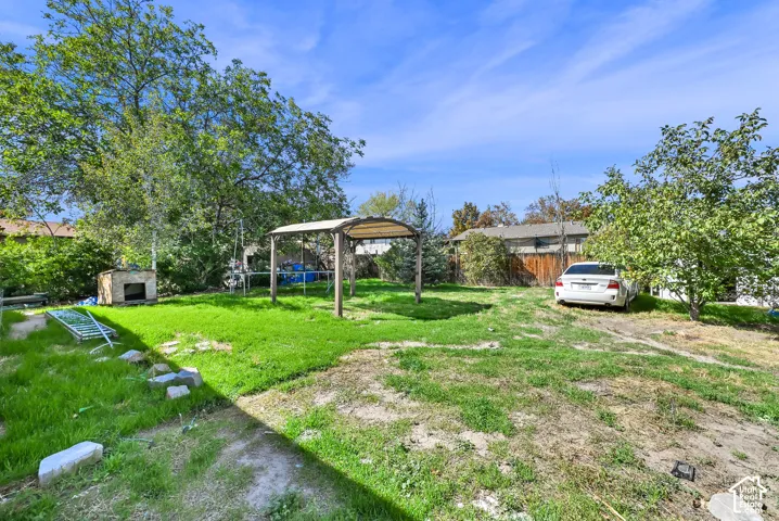 Expansive backyard featuring a partial grass lawn, mature trees, a built-in outdoor fireplace, and a standalone pergola