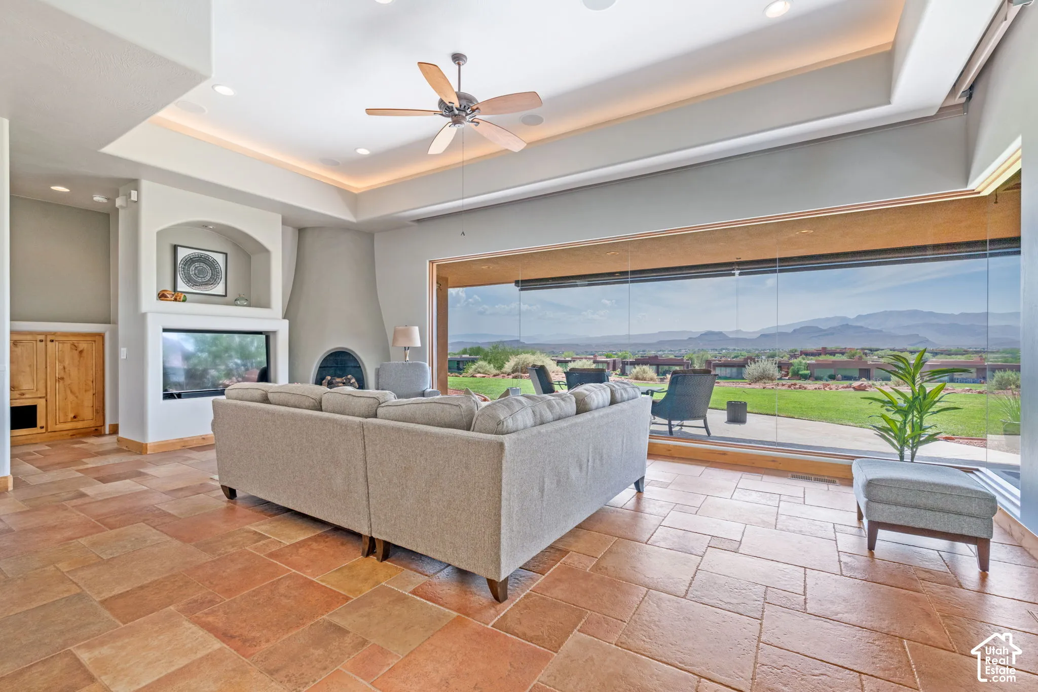 Expansive glass wall overlooking mountains, integrated fireplace, arched built-in shelving, tray ceiling with recessed lighting, and large format floor tile