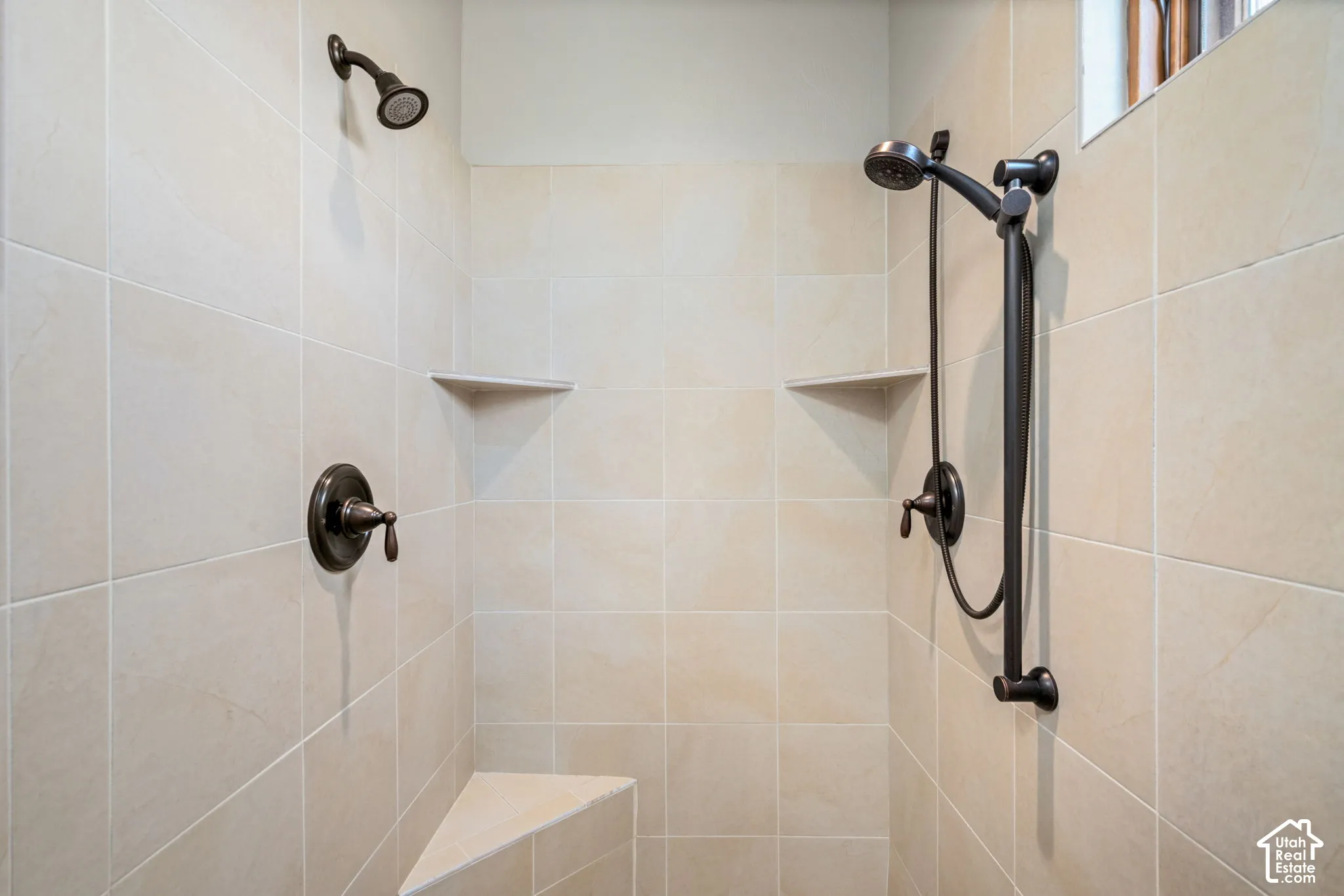 Tiled shower enclosure featuring a fixed showerhead, a handheld shower fixture, two built-in corner shelves, a built-in corner bench, and bronze-finish plumbing fixtures