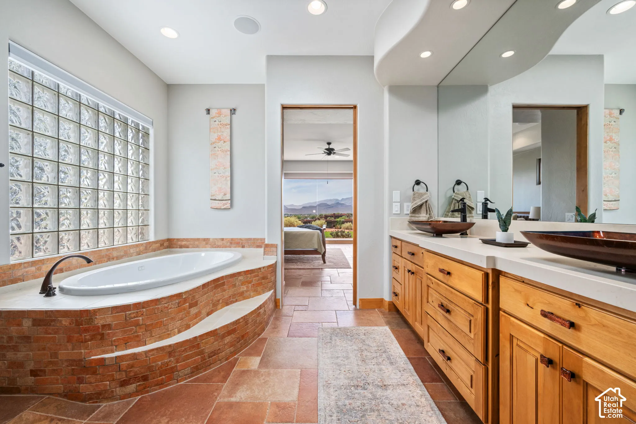 Soaking tub with a brick-finish surround, glass block window, dual vessel sinks, wood cabinetry, and extensive countertop space