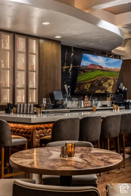 Illuminated display cabinetry, wood-paneled walls, a large wall-mounted screen, a stone-front bar with a light-colored countertop, and a round table with a polished stone-finish top