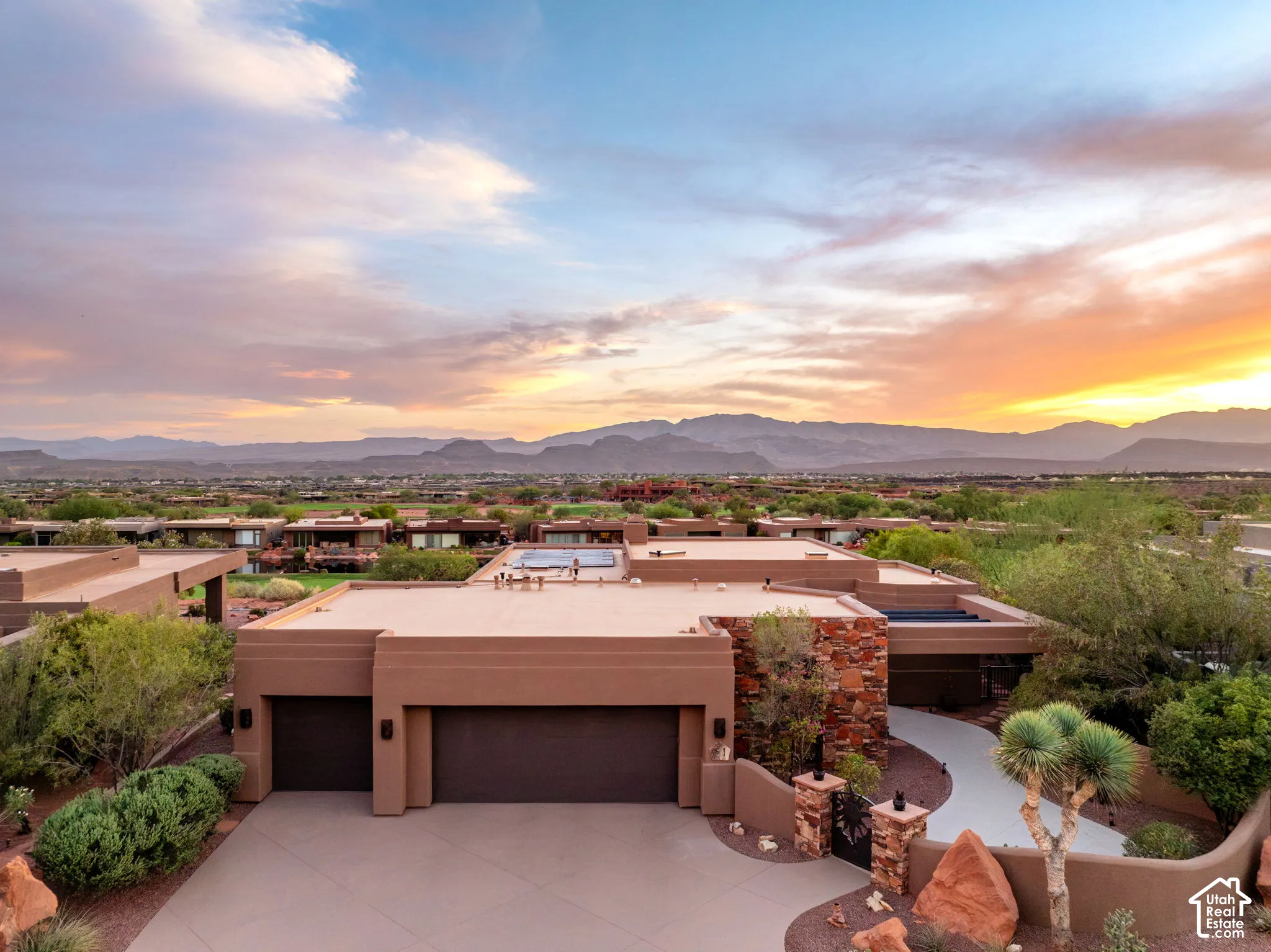 Terraced desert landscaping with mature plantings, stone exterior accents, and a paved driveway leading to integrated garages