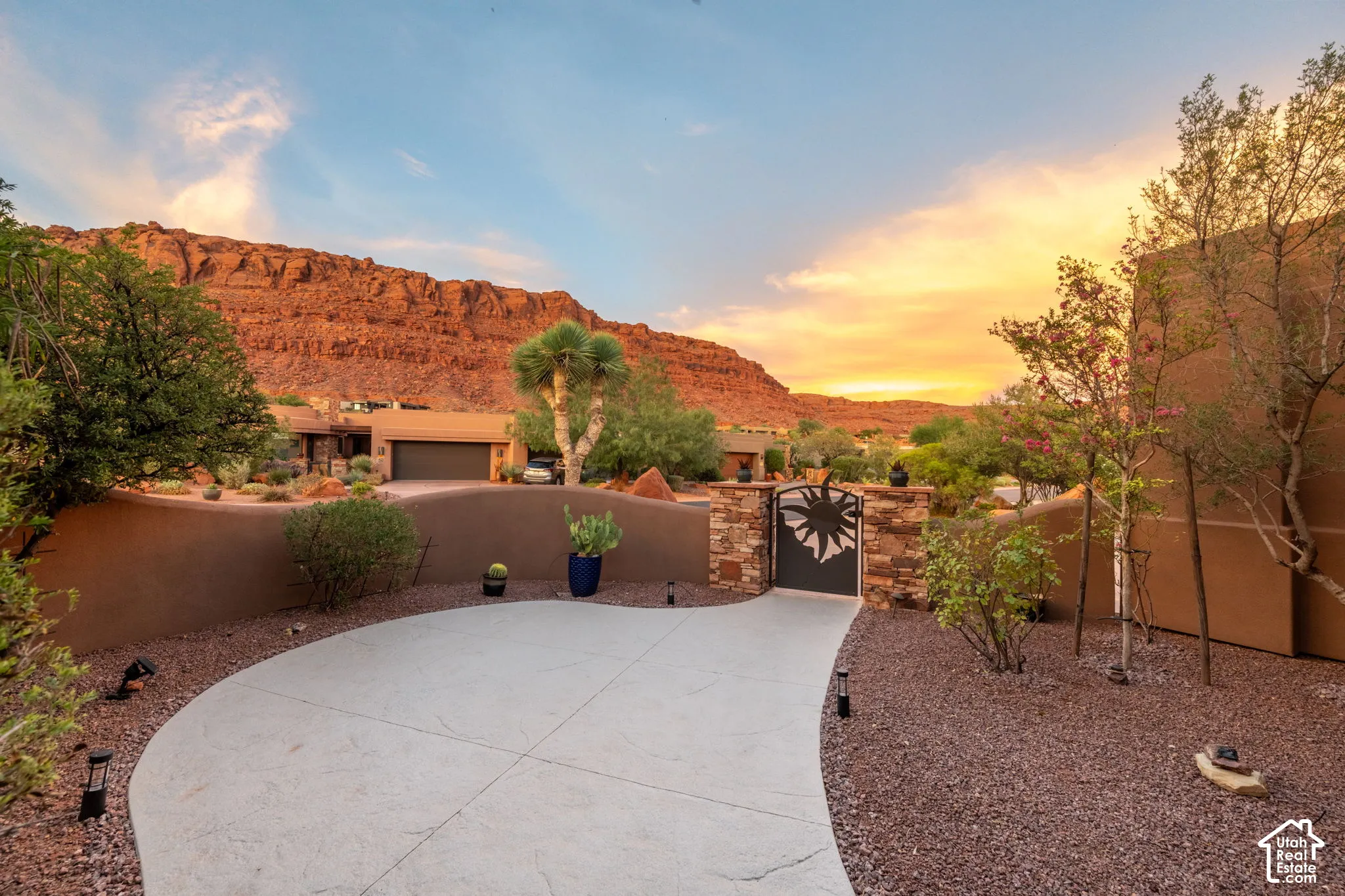 Curved concrete driveway with a decorative metal gate, stone column accents, and mature desert landscaping