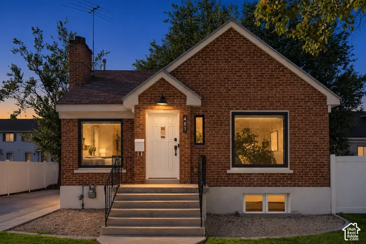 Brick exterior with a prominent chimney, concrete front steps with metal railings, a white front door with an overhead sconce, and a white picket fence