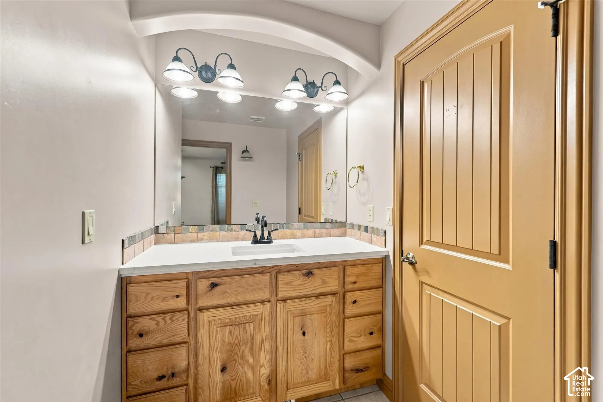 Vanity featuring natural-finish wood cabinetry, a solid-surface countertop with an integrated rectangular sink, a multi-light wall fixture, and a recessed arch architectural detail