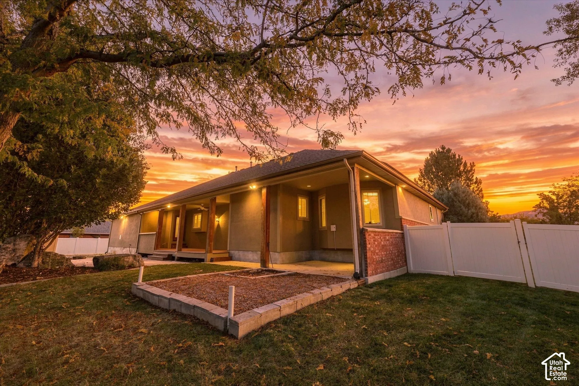 Stucco and brick exterior featuring a covered patio, raised stone-bordered landscaping bed, and white vinyl perimeter fencing