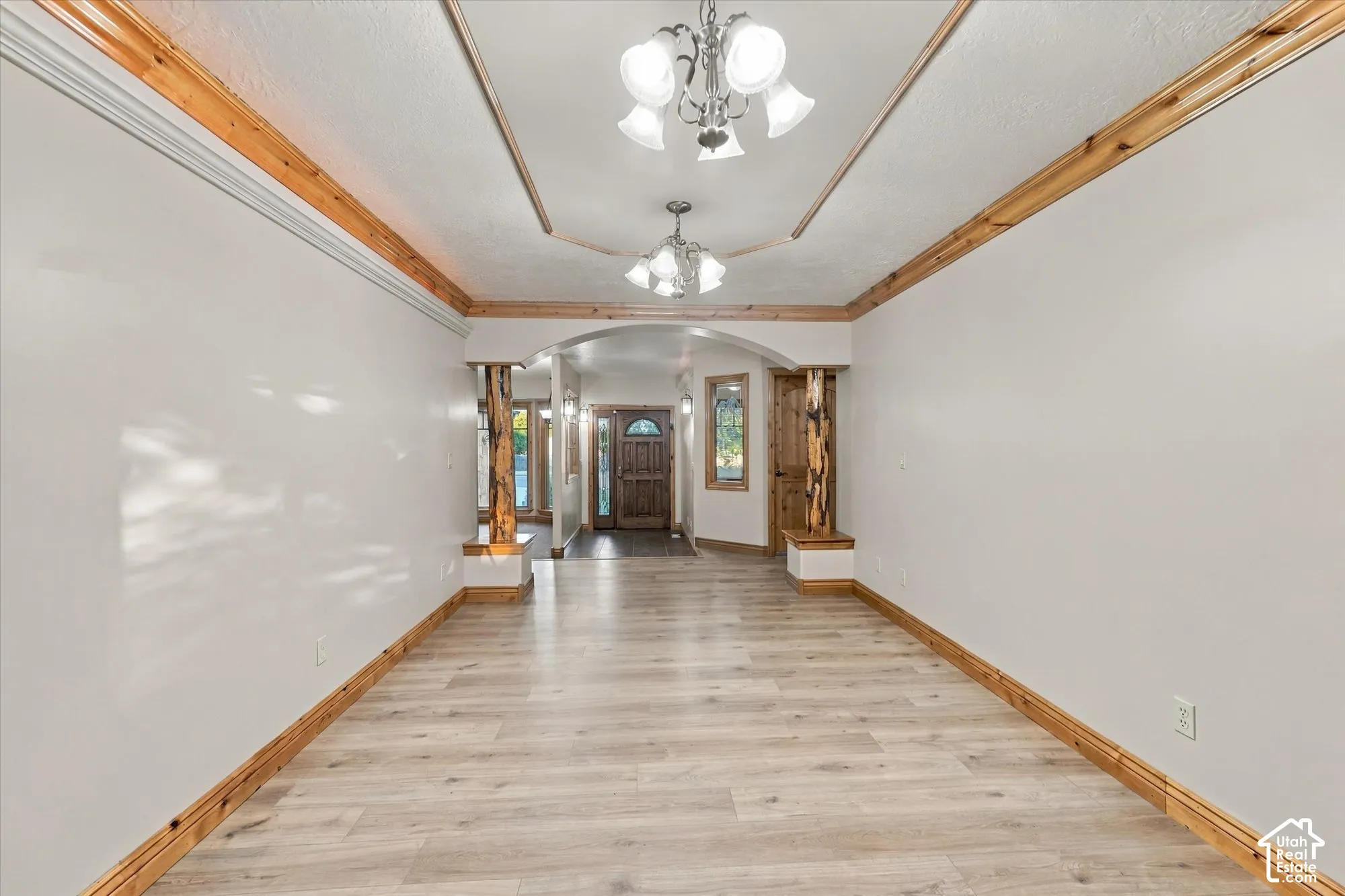 Interior space featuring wood-finish flooring, natural wood crown molding, a tray ceiling with dual multi-arm chandeliers, and decorative wood pillars