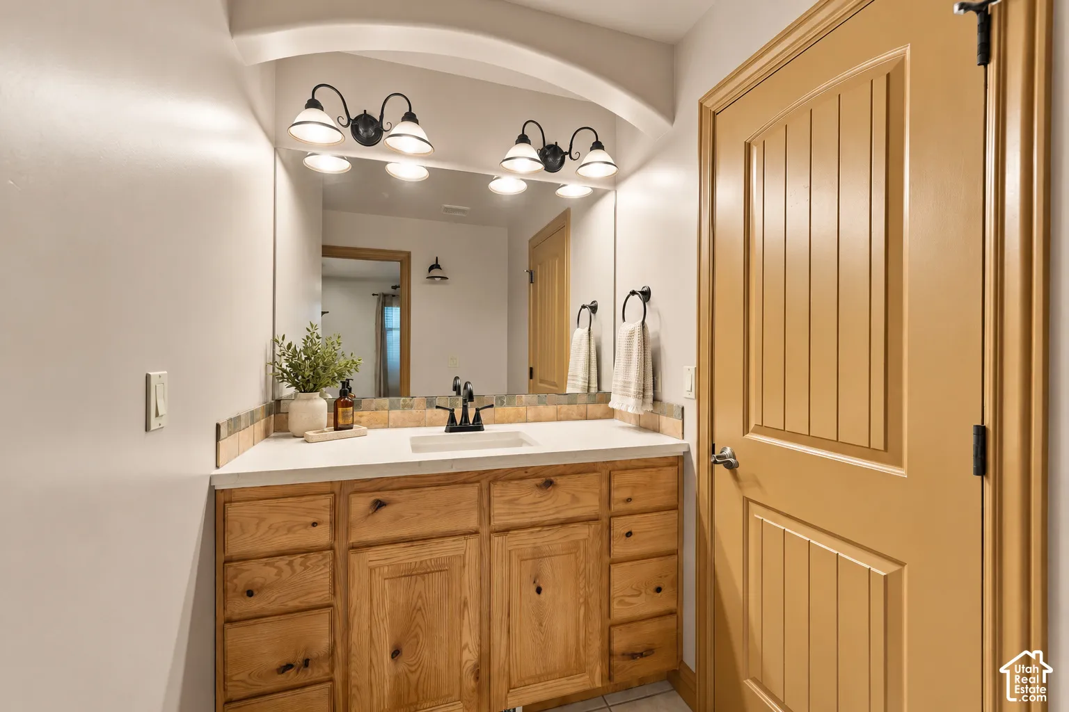 Bathroom vanity featuring a light-finish wood cabinet, white countertop, integrated rectangular sink, oil-rubbed bronze faucet, and dual-light wall sconces beneath a recessed arched alcove