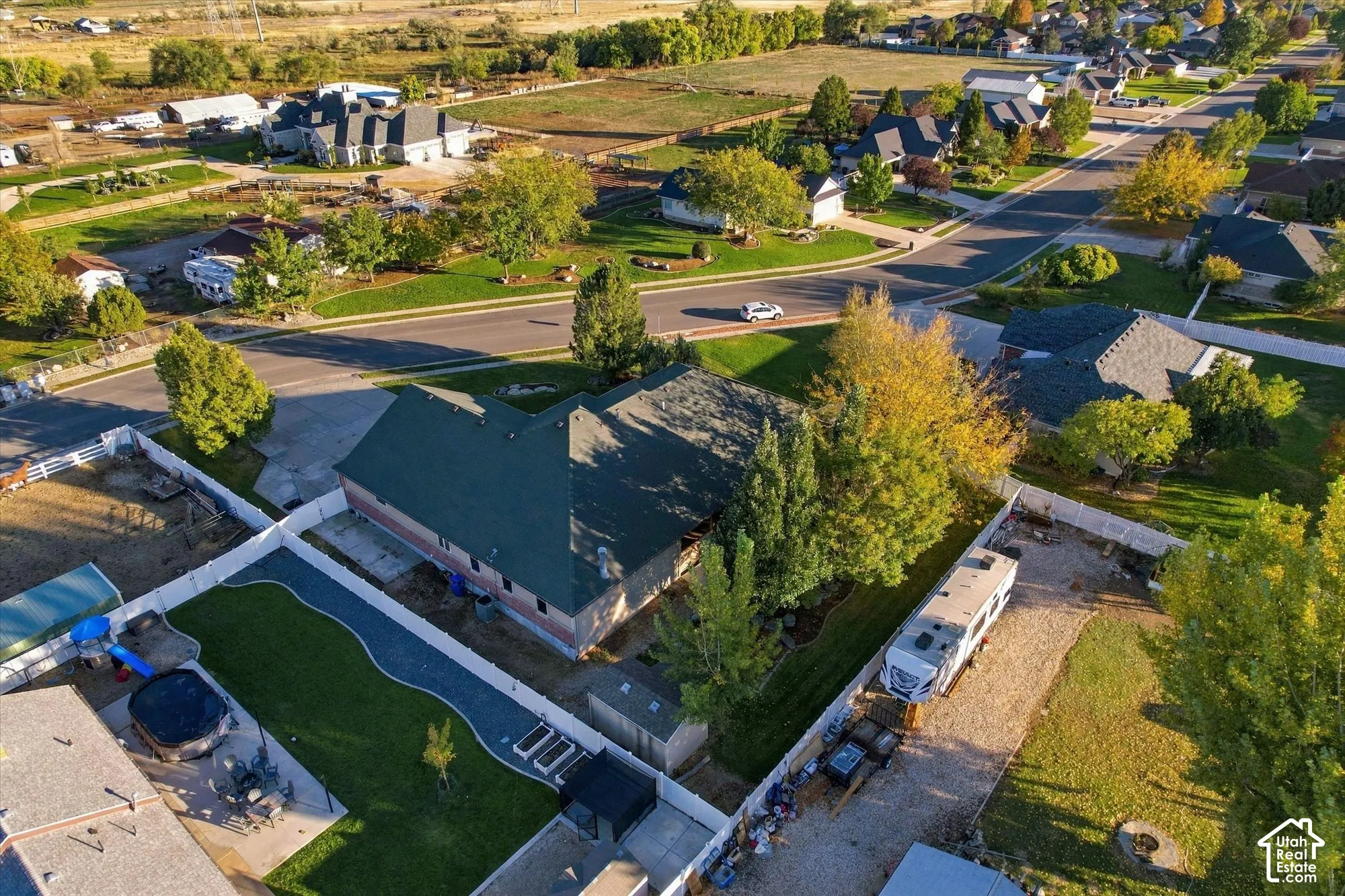 Aerial perspective featuring a residential lot with a fenced backyard, integrated concrete patio, mature trees, and a dedicated gravel side access area