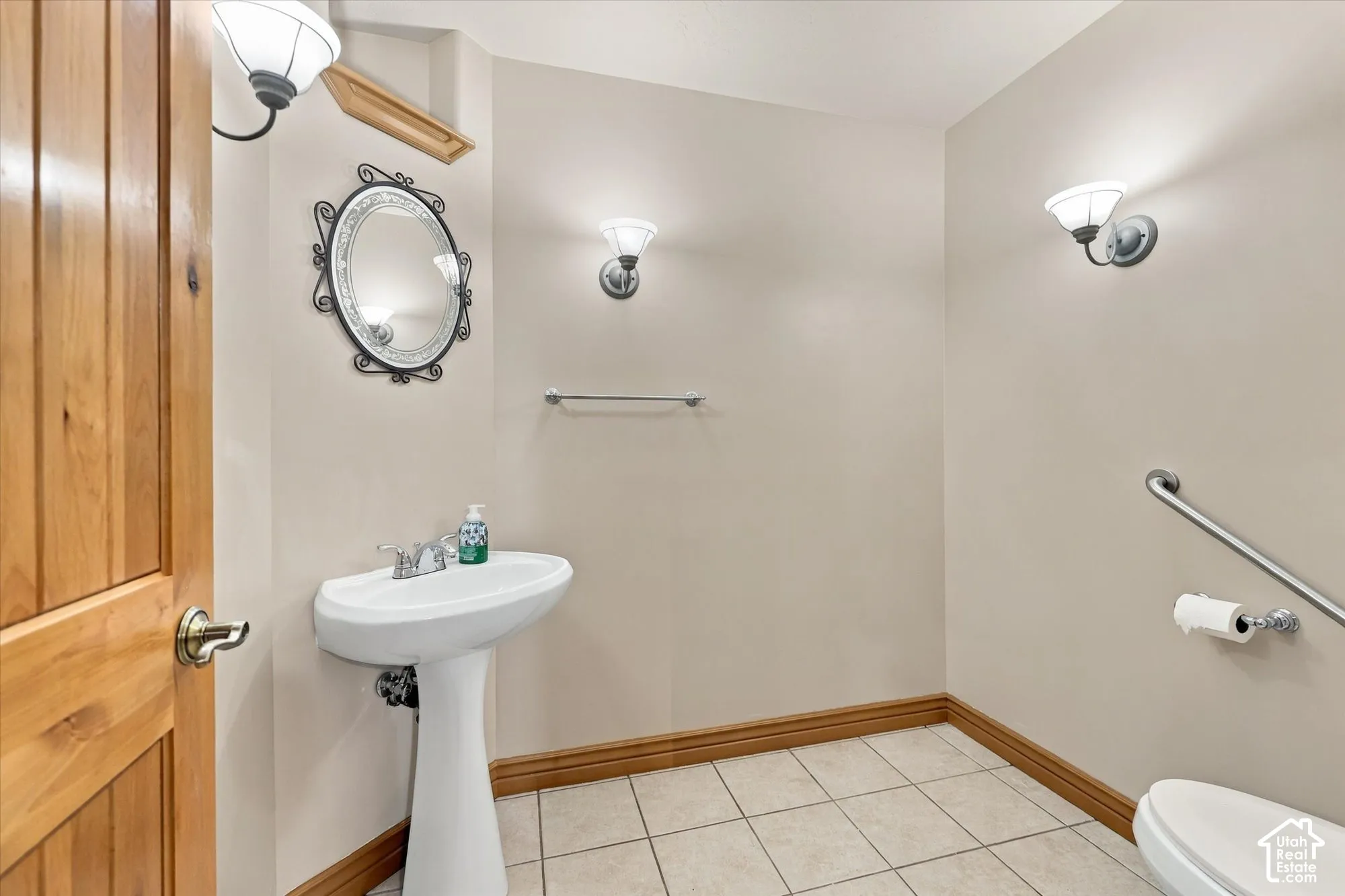Pedestal sink with chrome faucet, ceramic tile flooring, neutral wall finish, polished wood-finish door, and wall-mounted light fixtures