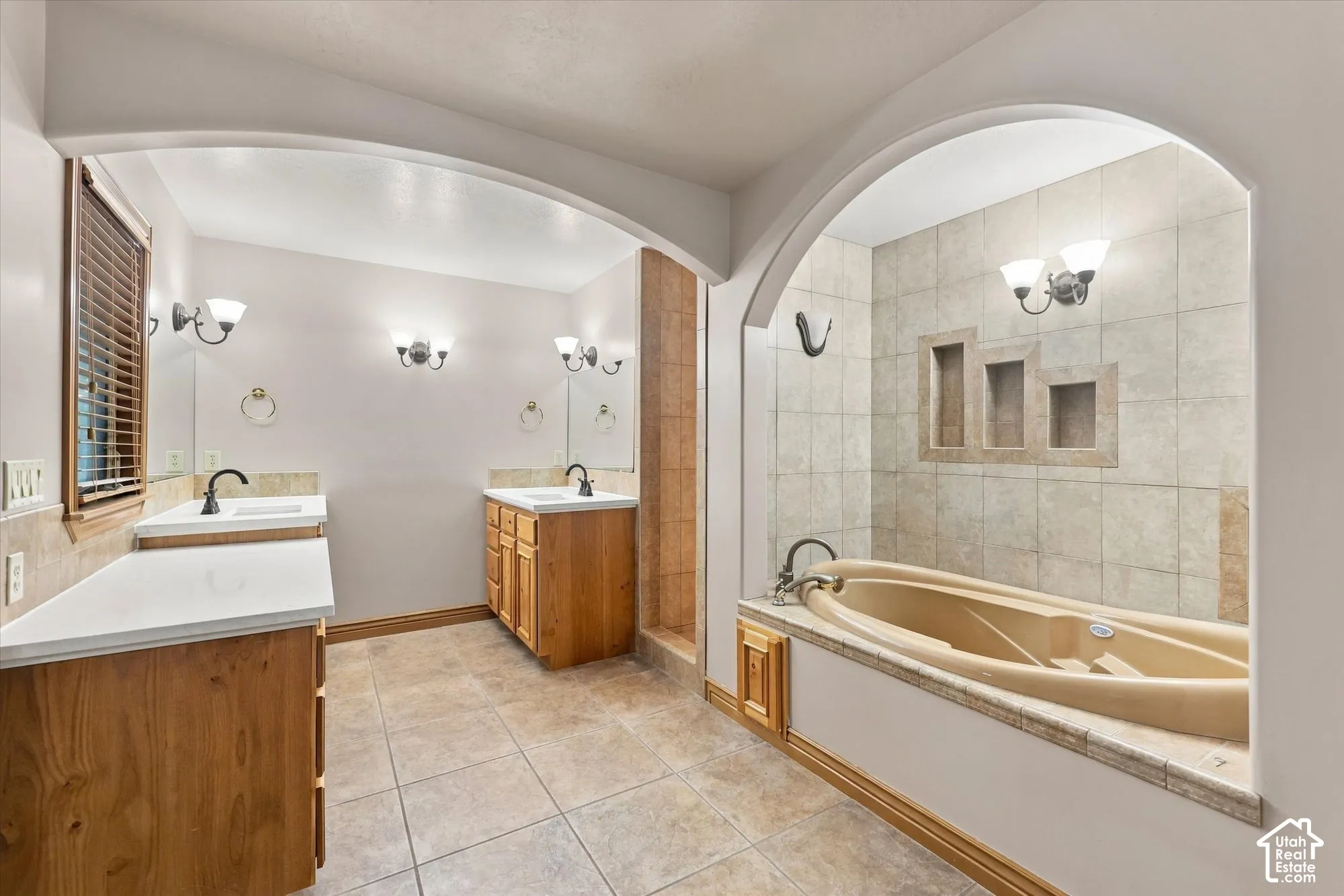Dual vanities featuring wood cabinetry and white countertops, tile flooring, arched alcove with a jetted soaking tub, tiled shower surround with built-in niches, and wall-mounted light fixtures