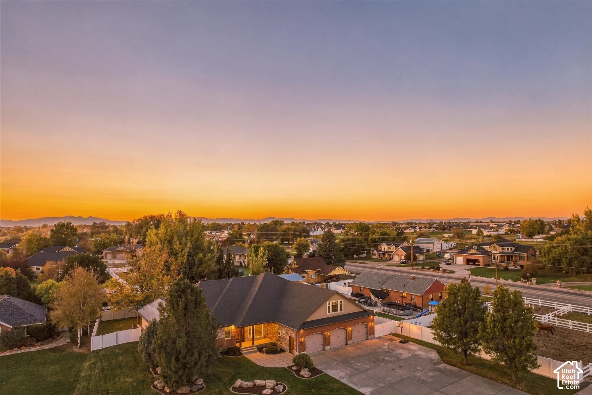 Aerial perspective of a multi-gabled residence featuring a three-car garage, stone masonry accents, paved driveway, and white vinyl perimeter fencing