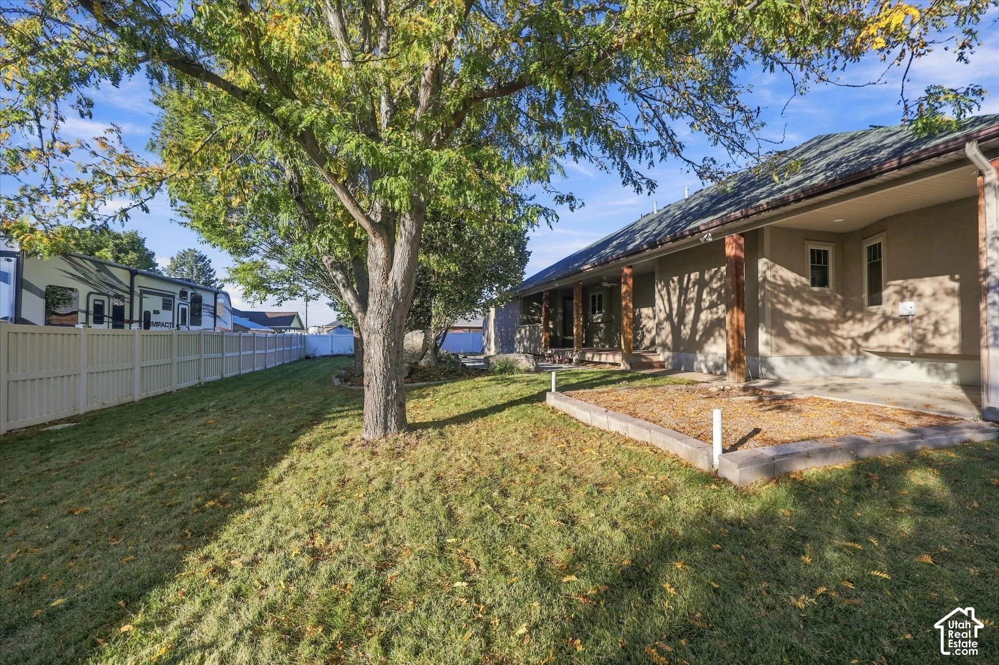 Backyard featuring a concrete covered patio, raised garden bed with masonry borders, mature trees, and perimeter vinyl fencing