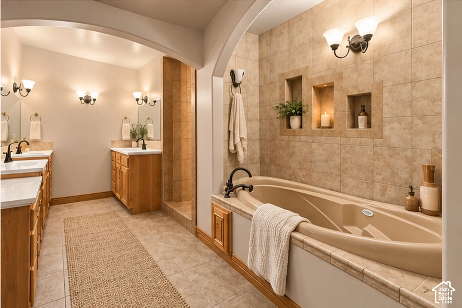 Bathroom featuring dual wood-finish vanities with rectangular sinks, a deep soaking tub with tile surround, arched architectural detailing, and recessed wall shelving