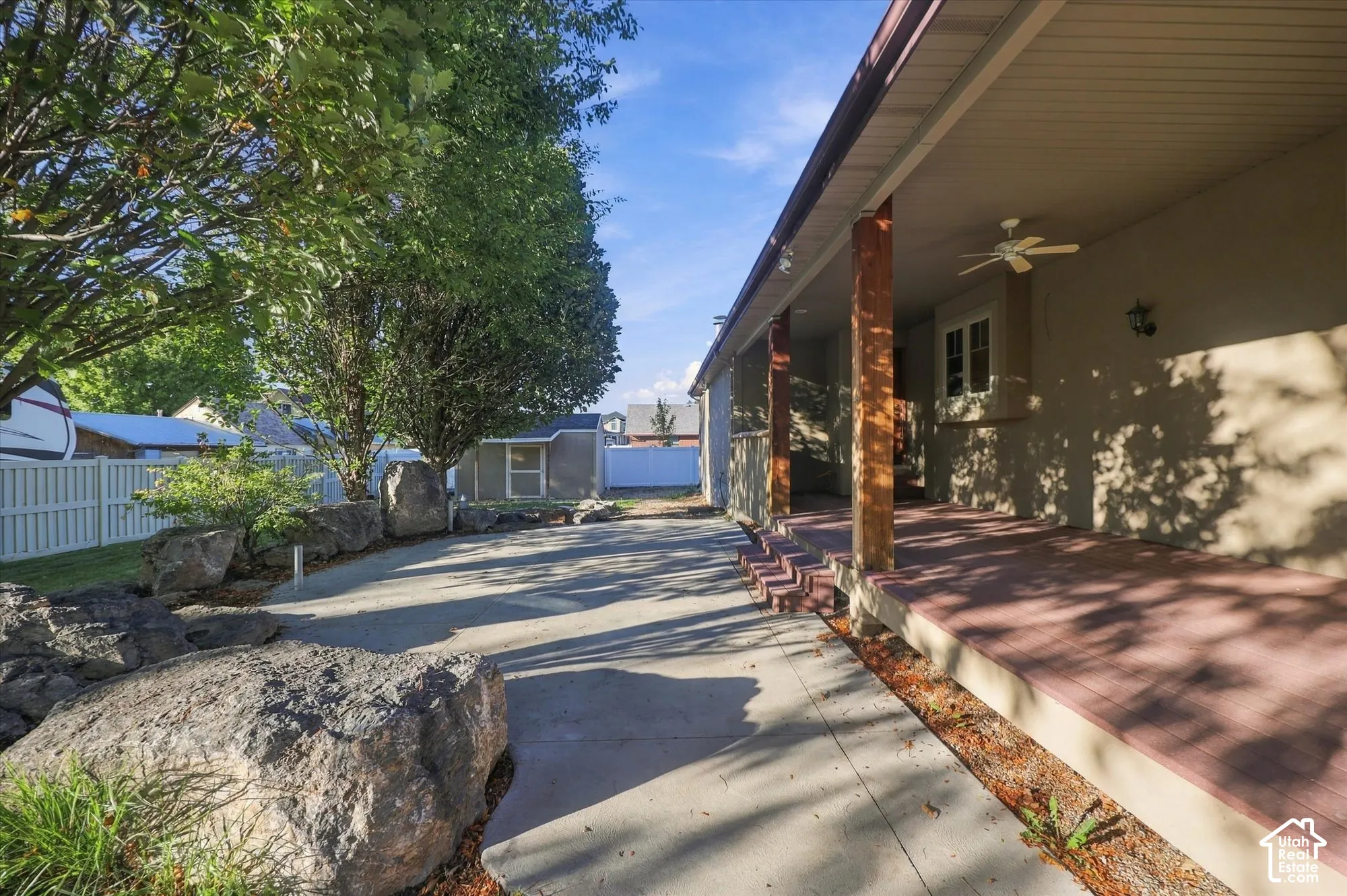 Covered patio featuring wood-finished posts, a ceiling-mounted fan, concrete hardscaping, large decorative boulders, and white vinyl privacy fencing