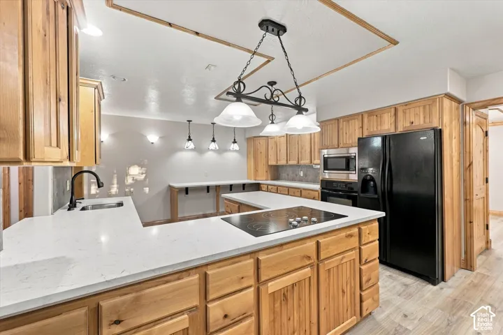 Kitchen featuring natural wood cabinetry, light-toned stone-finish countertops, an integrated glass cooktop, built-in black appliances, and decorative tiered pendant lighting