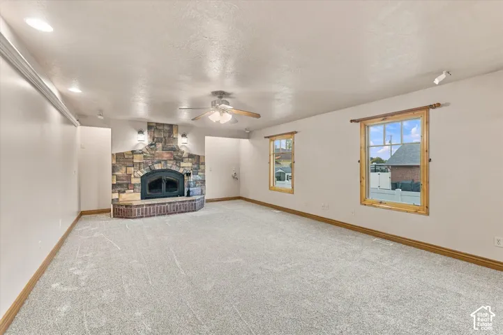 Stone fireplace with a raised brick hearth, neutral wall-to-wall carpeting, recessed lighting, ceiling fan, and wood-trimmed windows