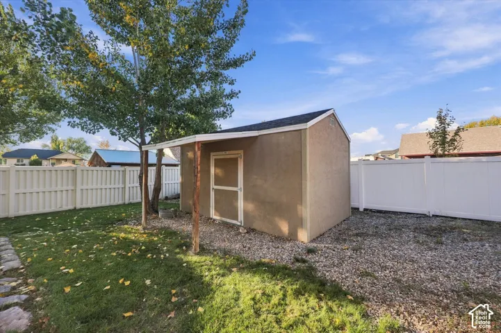 Stucco storage shed featuring a pitched shingle roof, a covered entry with a wooden support post, and a surrounding white vinyl privacy fence