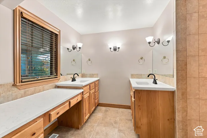 Dual vanity featuring light wood-finish cabinetry, solid surface countertops with integrated sinks, tile backsplash, and wall-mounted light fixtures