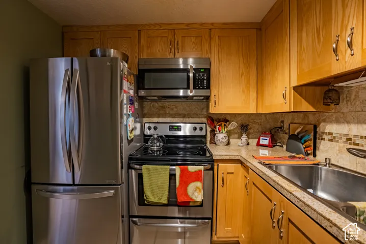 Wood-finish cabinetry, stainless steel appliances, stone-look countertops, tile backsplash, and stainless steel sink