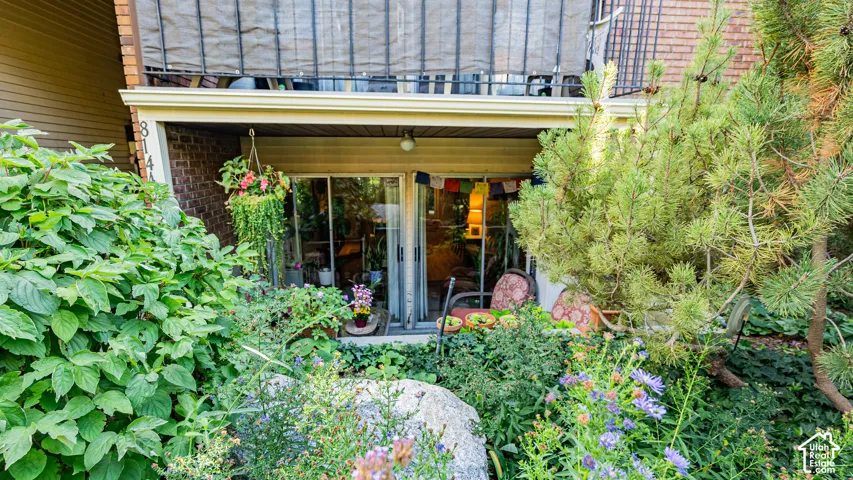 Ground-level patio with sliding glass doors, brick exterior accents, and diverse landscaping featuring shrubs and flowering plants