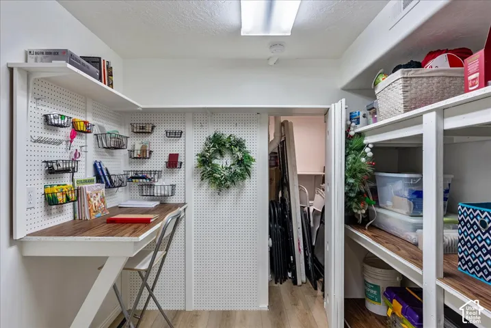 Built-in desk workspace featuring wall-mounted pegboard organization and wood-finish surfacing, accompanied by floor-to-ceiling stationary shelving and overhead fluorescent lighting