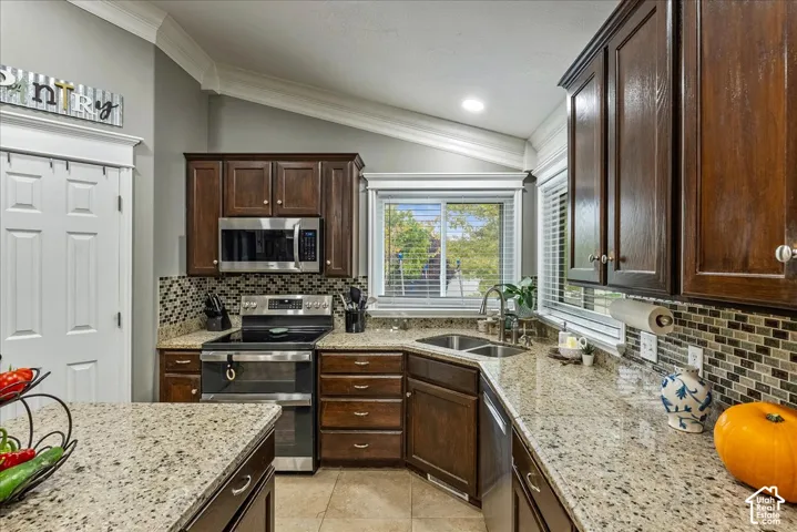 Kitchen featuring rich wood cabinetry, stone-finish countertops, decorative tile backsplash, stainless steel appliances, and vaulted ceilings with crown molding