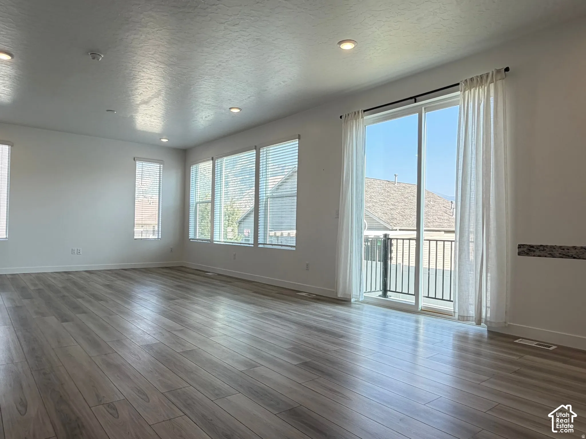 Spacious interior with wood-finish flooring, recessed lighting, multiple windows with blinds, and a glass sliding door leading to a balcony with black metal railings