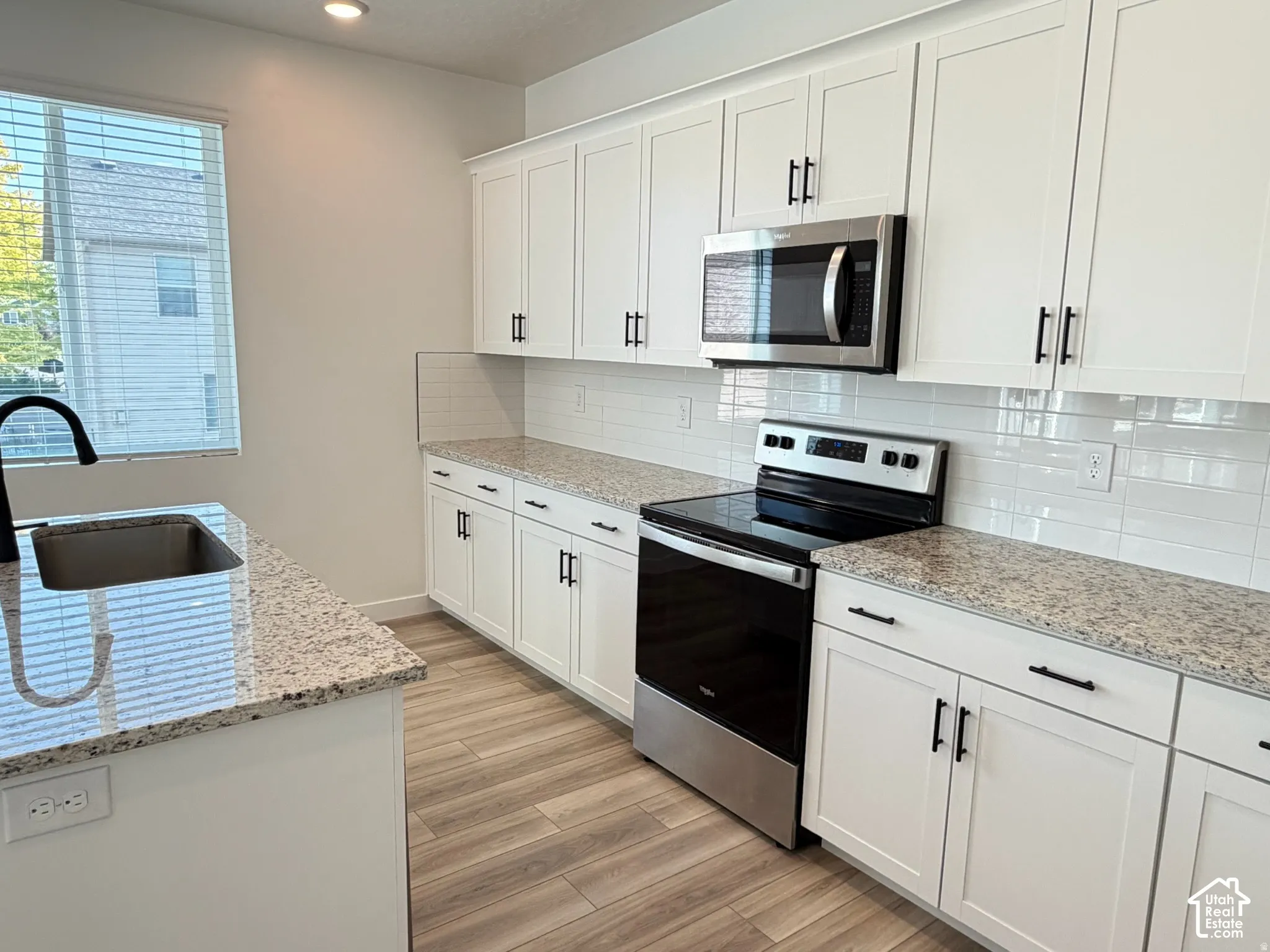White shaker-style cabinetry with black hardware, stainless steel appliances, granite countertops, subway tile backsplash, and wood-finish plank flooring