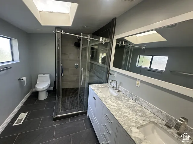 Dual vanity with stone countertop, frameless shower enclosure, skylight, recessed lighting, and dark tile flooring