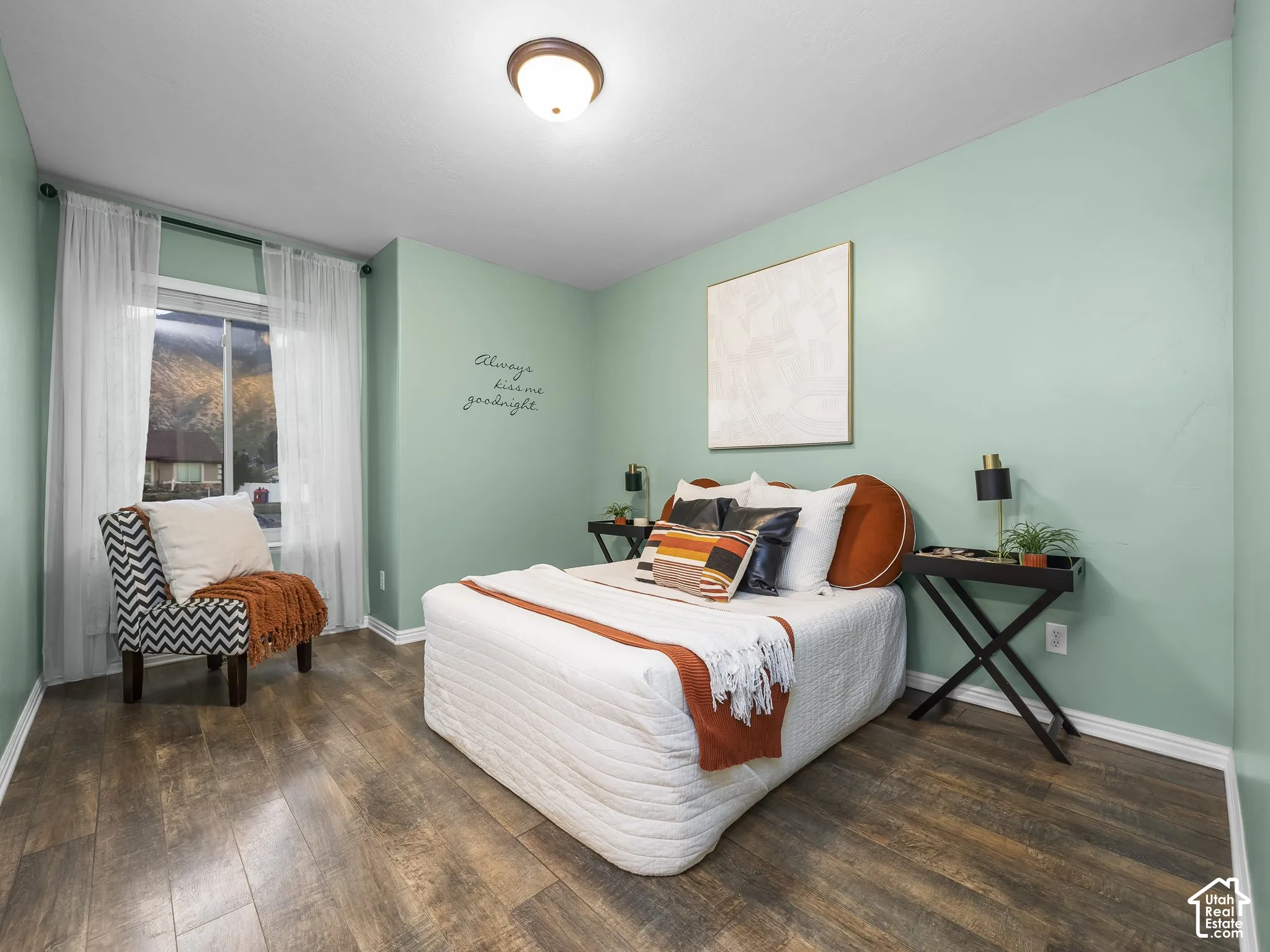 Serene bedroom retreat featuring soft sage-green walls, wood-finish flooring throughout, and abundant natural light from a generous window with sheer curtains
