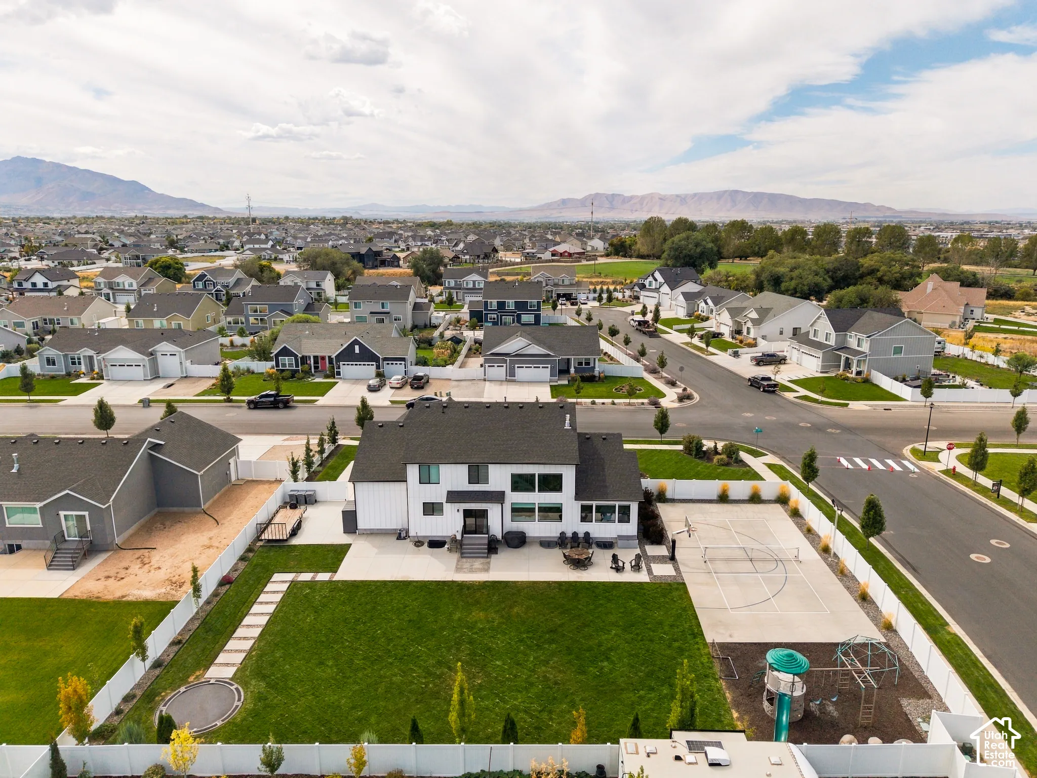 Two-story residence featuring a white vertical siding exterior, an expansive backyard with a manicured lawn, a paved sport court, and a separate play area