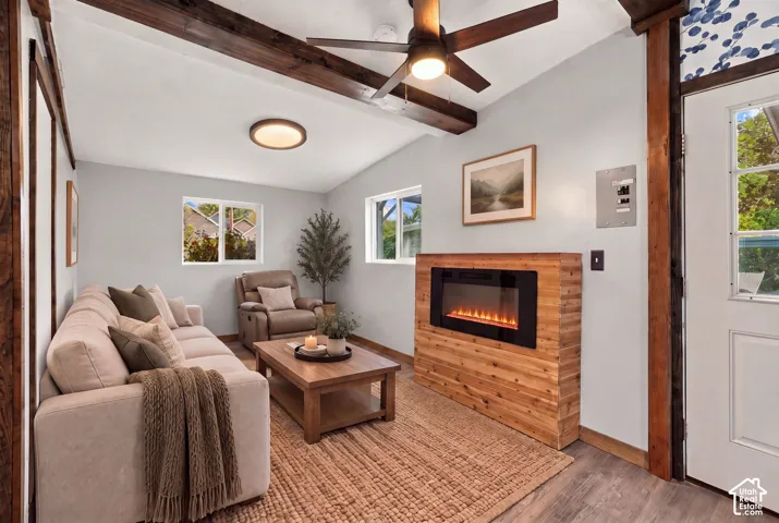 Vaulted ceiling with exposed wood beams, wood-finish flooring, built-in electric fireplace with wood surround, ceiling fan with integrated lighting, and multiple windows