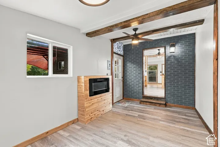 Wood-finish flooring, exposed wood beams, a built-in fireplace, a ceiling fan with integrated lighting, and an accent brick wall
