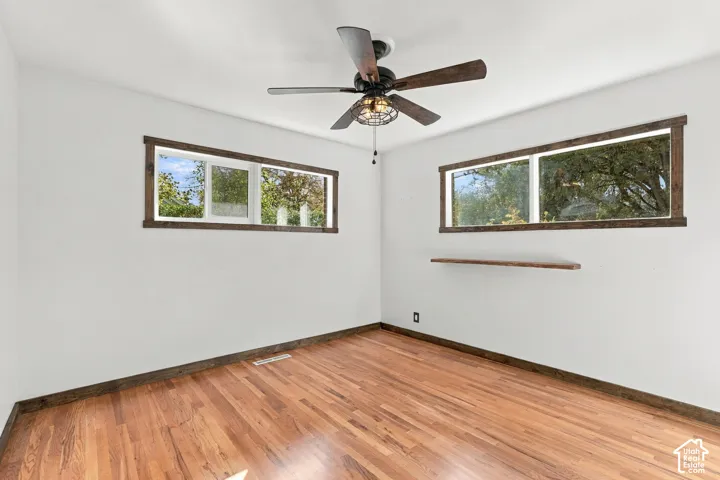 Hardwood flooring, two horizontal windows with wood trim, ceiling fan with light fixture, and a floating wood shelf
