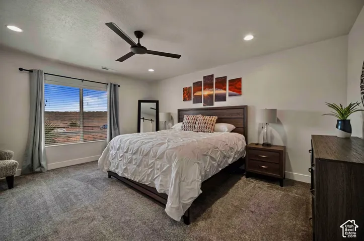Recessed ceiling lighting, ceiling fan, large window with blinds, wood-finish headboard, and gray carpeting