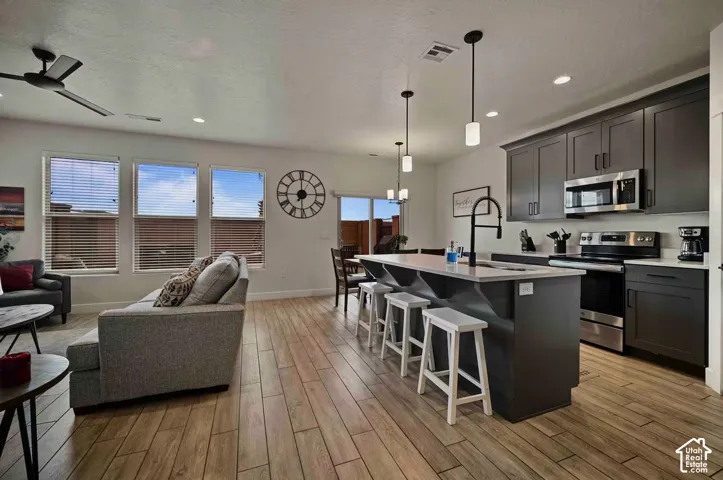 Open-concept living space featuring wood-finish flooring, a modern kitchen island with pendant lighting, dark cabinetry, and stainless steel appliances