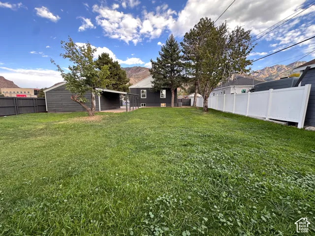 Expansive backyard featuring a lawn, mature evergreen trees, a covered outdoor structure, and white vinyl perimeter fencing, set against a mountain backdrop