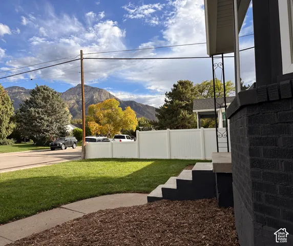 Exterior of the home featuring dark-painted brick, a concrete step entryway with decorative wrought iron supports, a lawn, mulch landscaping, and a solid white perimeter fence against a mountain backdrop