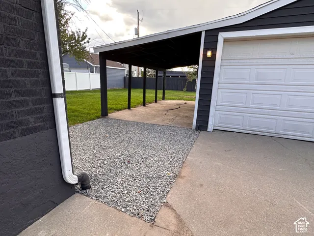 Attached garage with white panel door, covered carport, gravel ground covering, and a white privacy fence