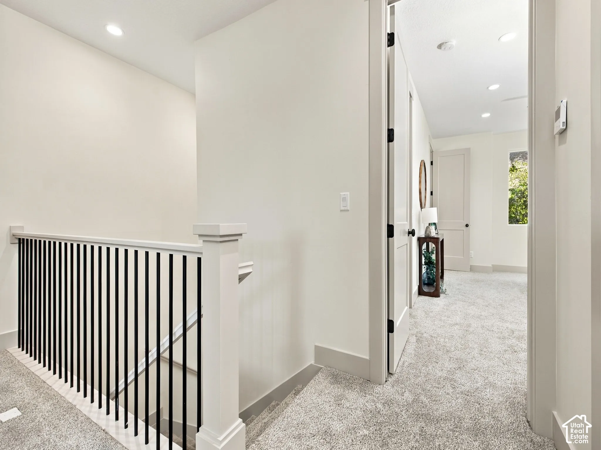 Staircase with black metal balusters and a white newel post, featuring light gray carpeting throughout the hallway and recessed ceiling lighting