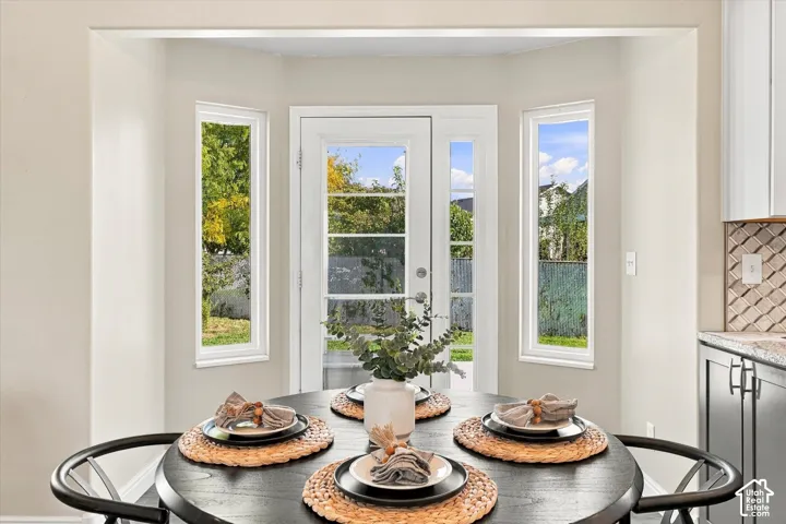 Dining area showcasing a round table with woven placemats and place settings, anchored by three tall windows with white frames offering natural light and outdoor scenery