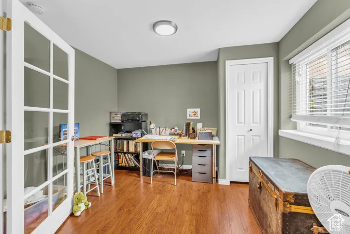 Versatile home office featuring warm wood-finish flooring, sage green walls, and abundant natural light from dual windows with plantation shutters